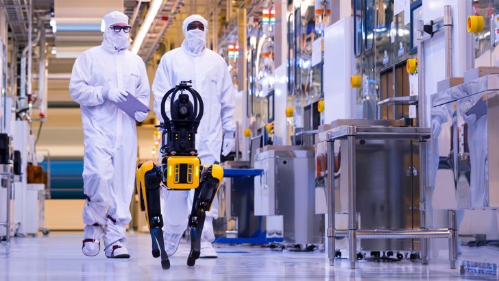 Two technicians in cleanroom suits walk alongside a yellow Boston Dynamics robot dog in a high-tech manufacturing facility.
