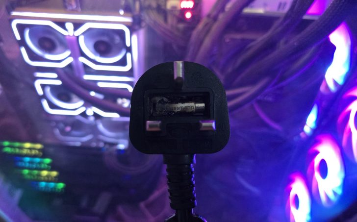 A black Bussmann plug with a visible fuse is held in front of a computer with illuminated RGB cooling fans and tubing.