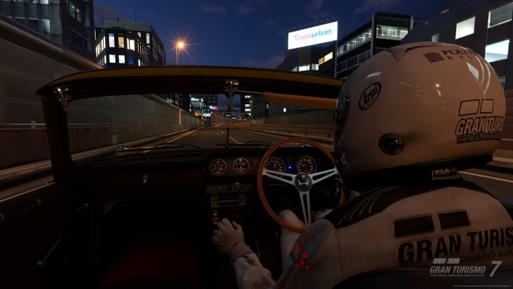 A driver wearing a Gran Turismo 7-branded helmet operates a classic convertible at night in an urban environment within the game Gran Turismo 7.