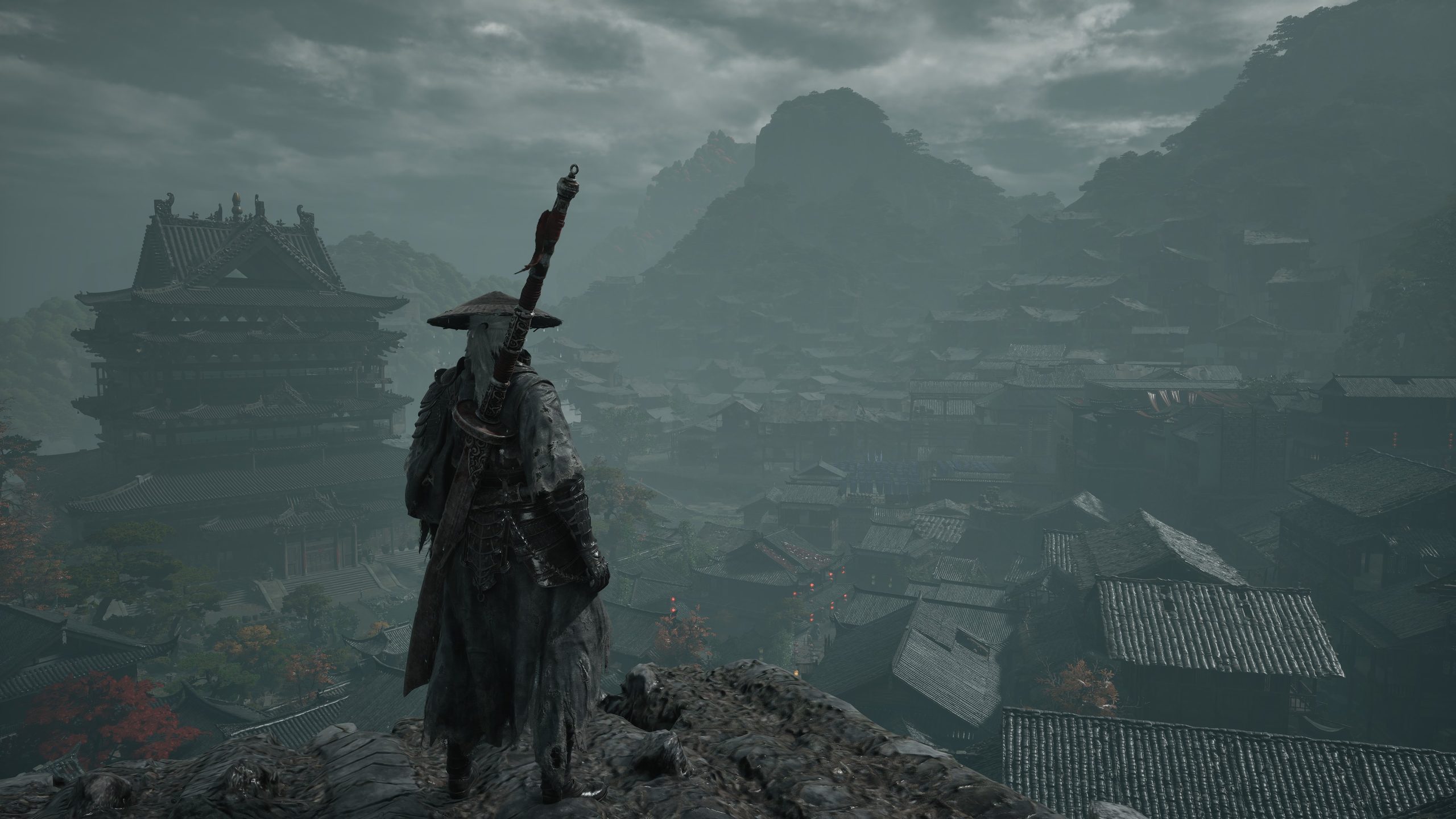 A character in traditional samurai attire stands on a rocky cliff overlooking a foggy, ancient Japanese village in a video game setting.