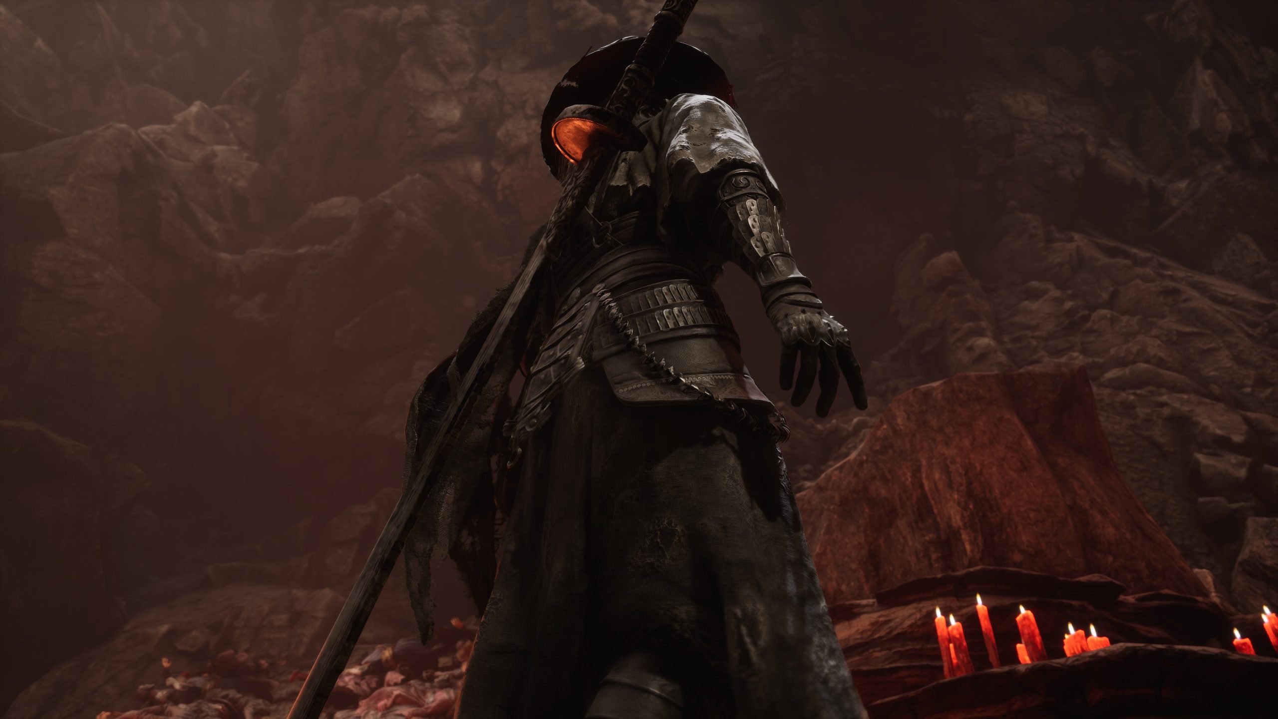 A character in dark armor, holding a sword, stands in a dimly lit cave with red candles on the ground in a scene from the game Lords of the Fallen.